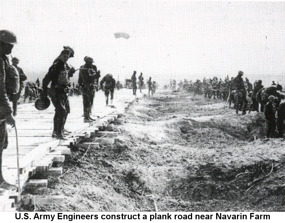 Black and white photo of army engineers wearing broad-brimmed flat World War I helmets, some holding tools, standing along the edge of a wide plank road supported by multiple layers of thick timbers and stones that stretches from the foreground into the distance; others work a few yards away in a long line in the ditch parallel to the road. An officer on horseback is visible in this second line.